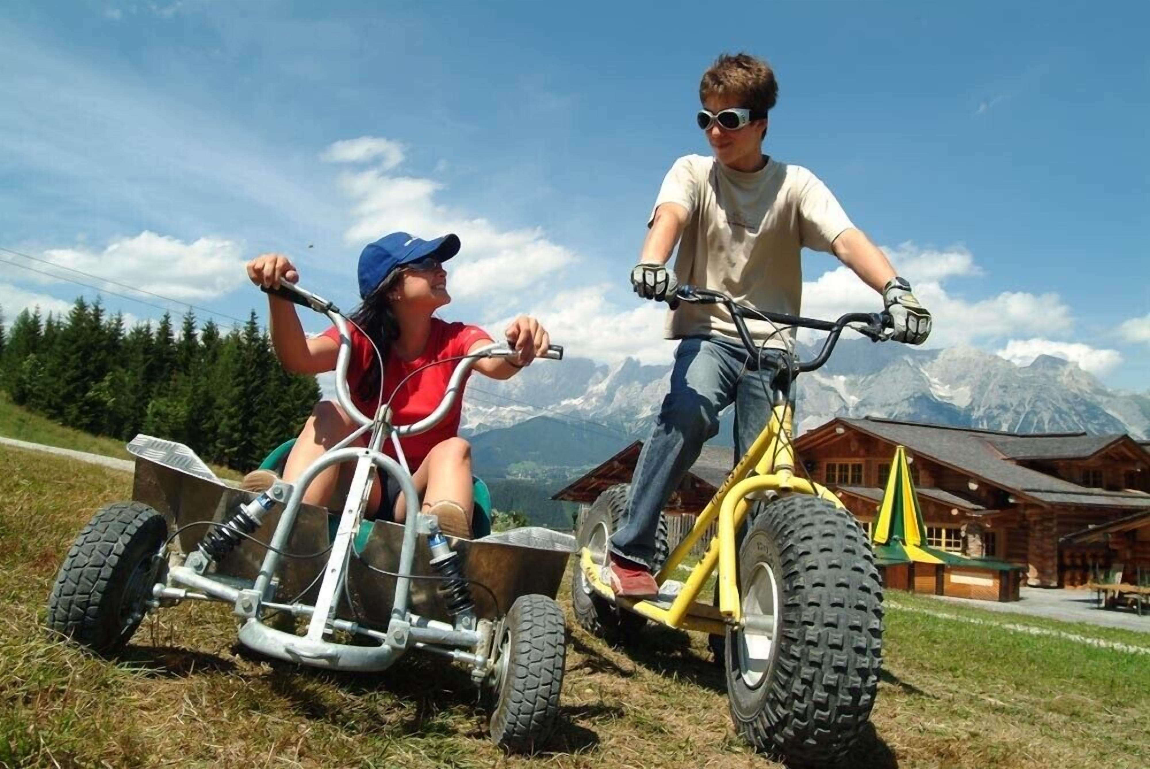 Deux personnes conduisent un petit véhicule tout-terrain sur un champ herbeux, avec des montagnes en arrière-plan.