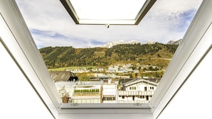 Vista desde una ventana con tragaluz, con vistas a un pueblo, edificios, una colina boscosa y montañas nevadas al fondo.