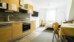 A modern kitchen with wooden cabinets, a built-in oven, and a microwave. There is a dining area with a table and chairs, and a bed with a wooden headboard.
