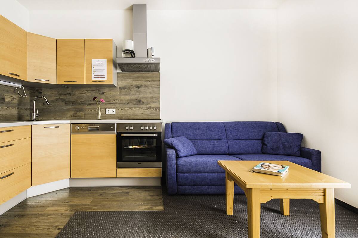 Une cuisine compacte avec des meubles en bois, un four à micro-ondes encastré et une cuisinière. On y trouve un canapé bleu et une petite table en bois sur laquelle est posé un magazine.