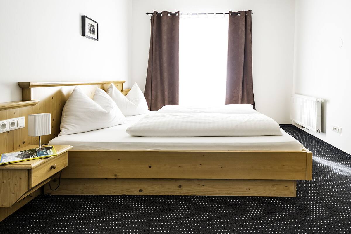 Un lit simple avec une tête de lit en bois, une table de chevet avec une lampe et un petit tiroir. Il y a une fenêtre avec des rideaux marron, un radiateur et un tableau au mur.