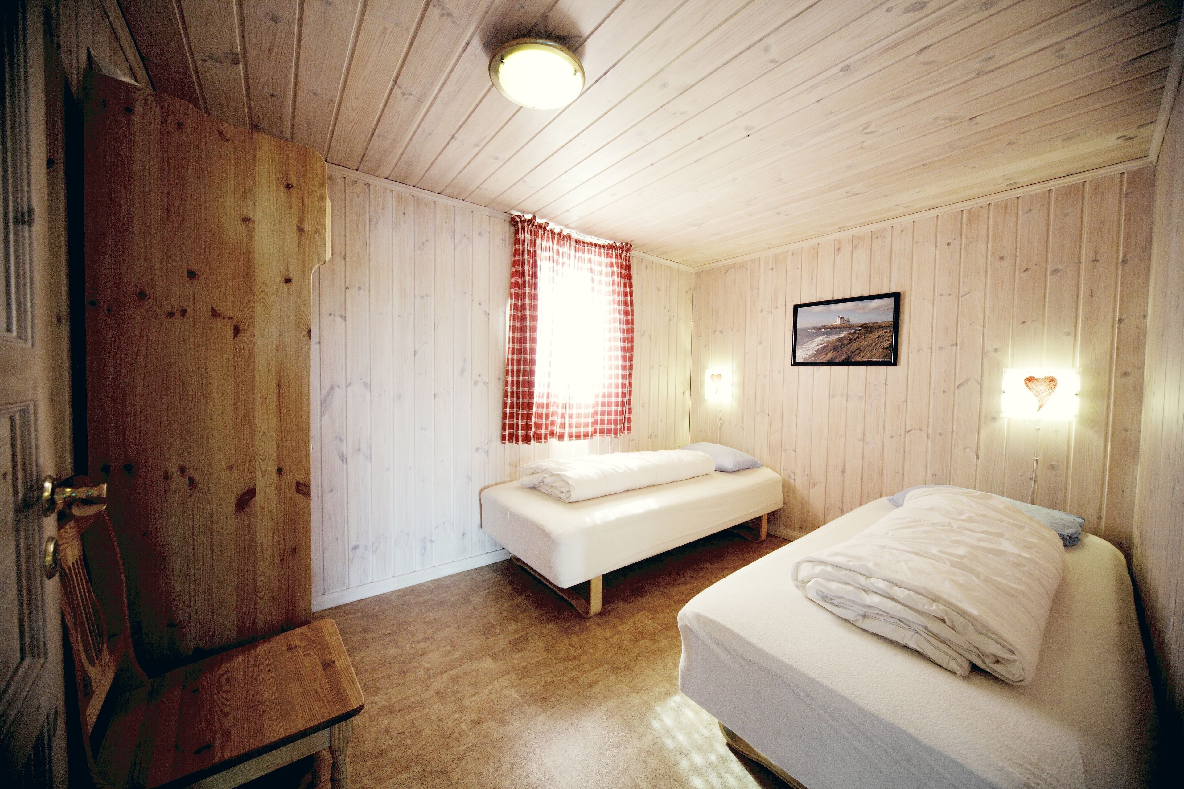 A room with wooden walls and floors, a bed with white bedding, a bench, and a window with red curtains.