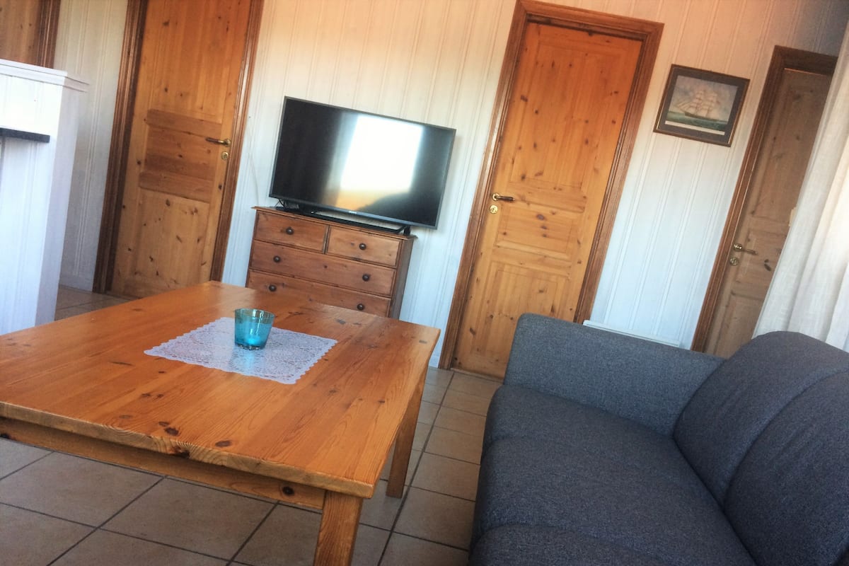 A wooden dining table with a blue glass and a lace placemat, a gray sofa, a television on a wooden cabinet, and wooden doors with a framed picture.