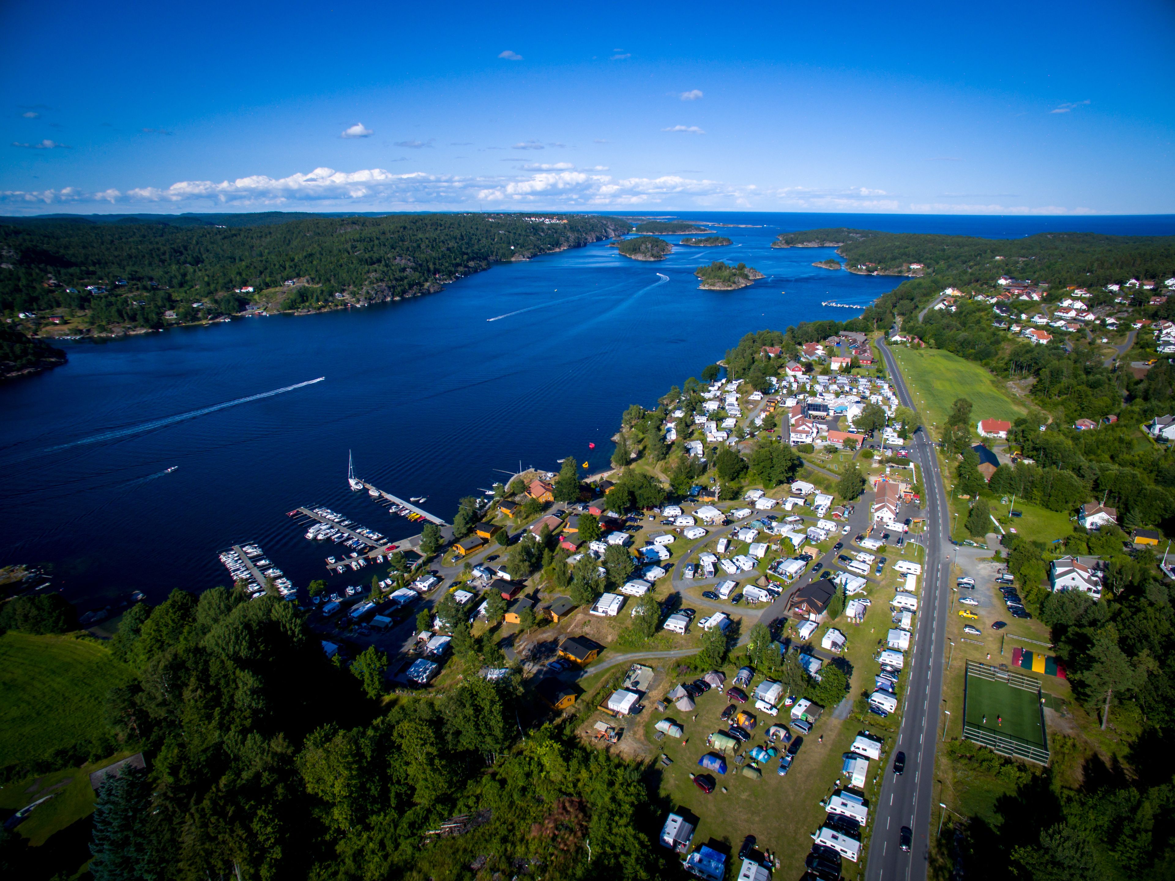 Vista aérea de un camping junto al lago con vehículos recreativos y tiendas de campaña, un puerto deportivo con barcos y una carretera que atraviesa la zona.