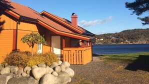 A wooden cabin with a red roof, situated by a lake with a view of a forested hill in the background. - Sørlandet Feriesenter (Risør)