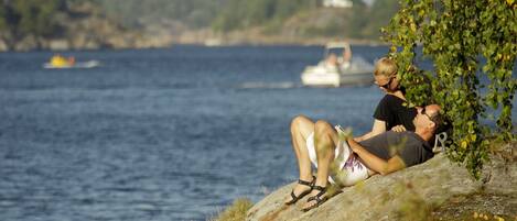 Una pareja descansando en una costa rocosa junto al agua, con barcos al fondo.