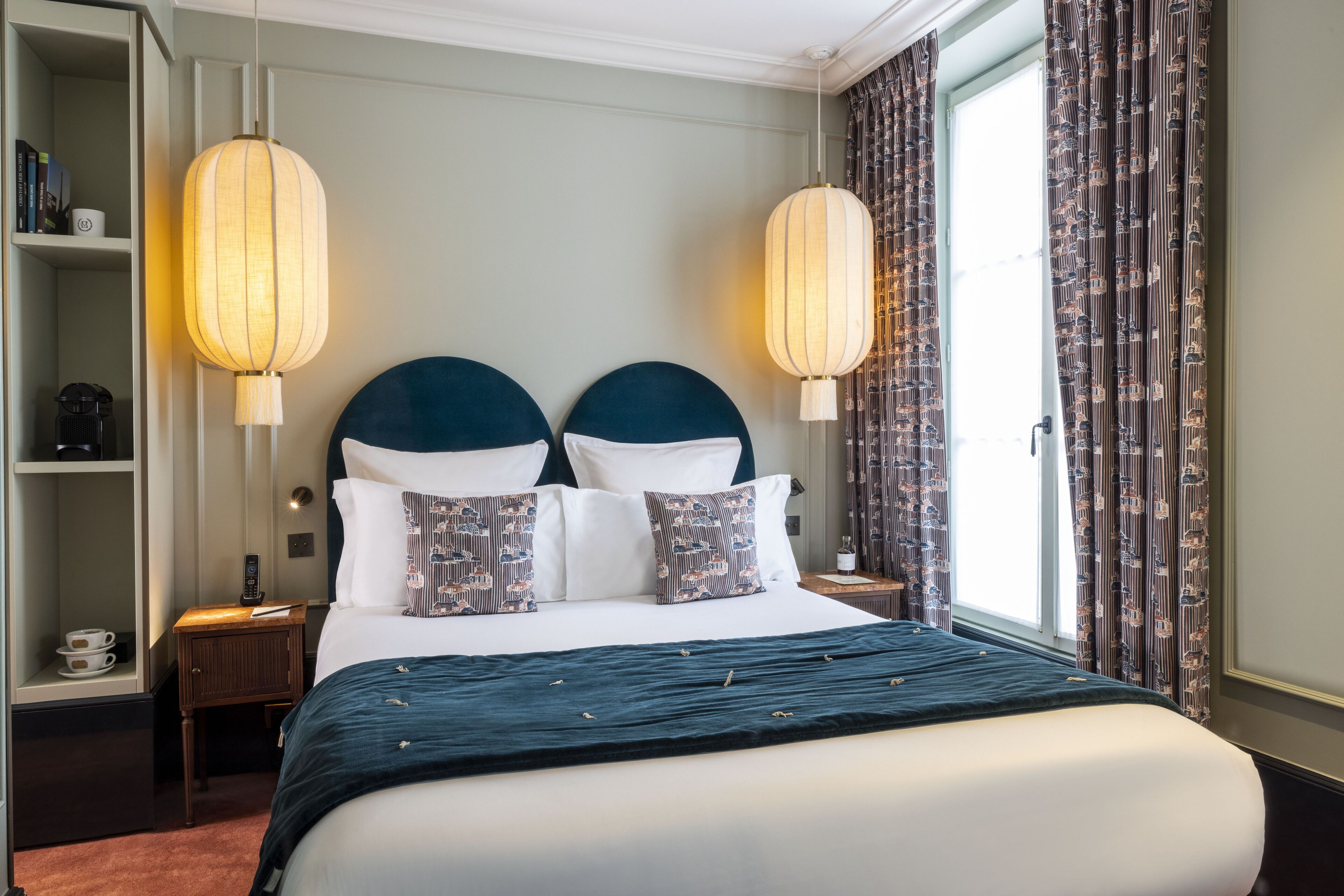 A hotel room with a large bed, two bedside lamps, a nightstand, a shelf with books and a coffee maker, and a window with curtains.