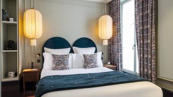 A hotel room with a large bed, two bedside lamps, a nightstand, a shelf with books and a coffee maker, and a window with curtains.