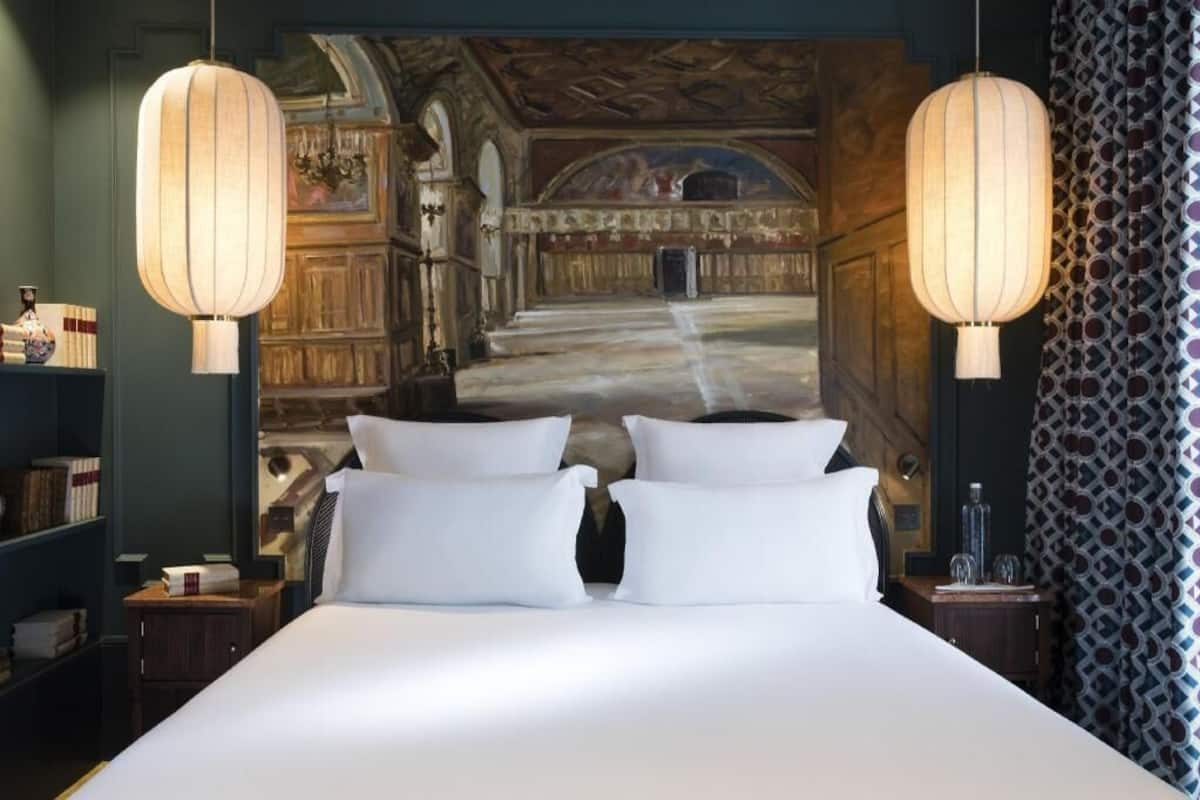A hotel room with a large bed, two bedside lamps, a bookshelf, and a mural on the wall.