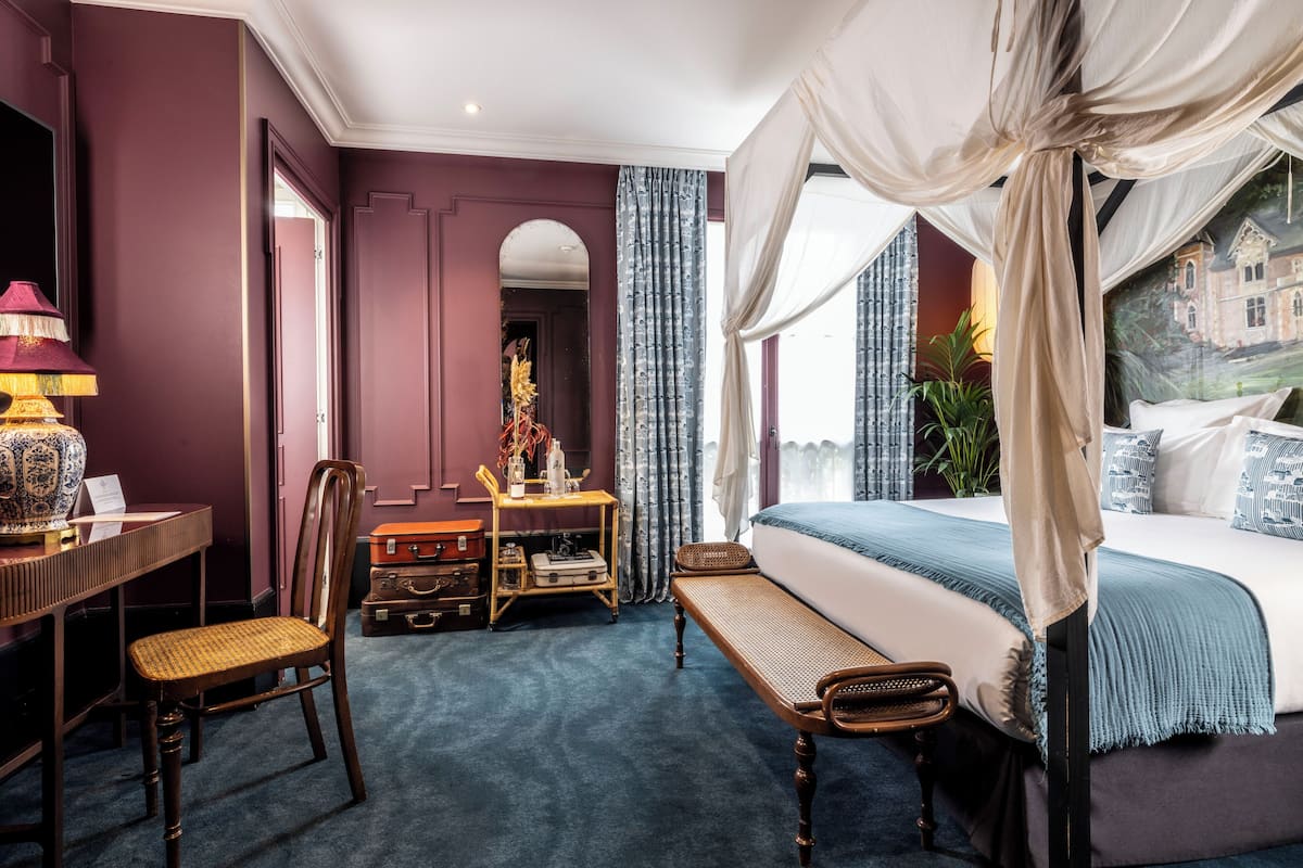 A bedroom with a large bed, a desk with a lamp, a chair, a bench, and a suitcase.