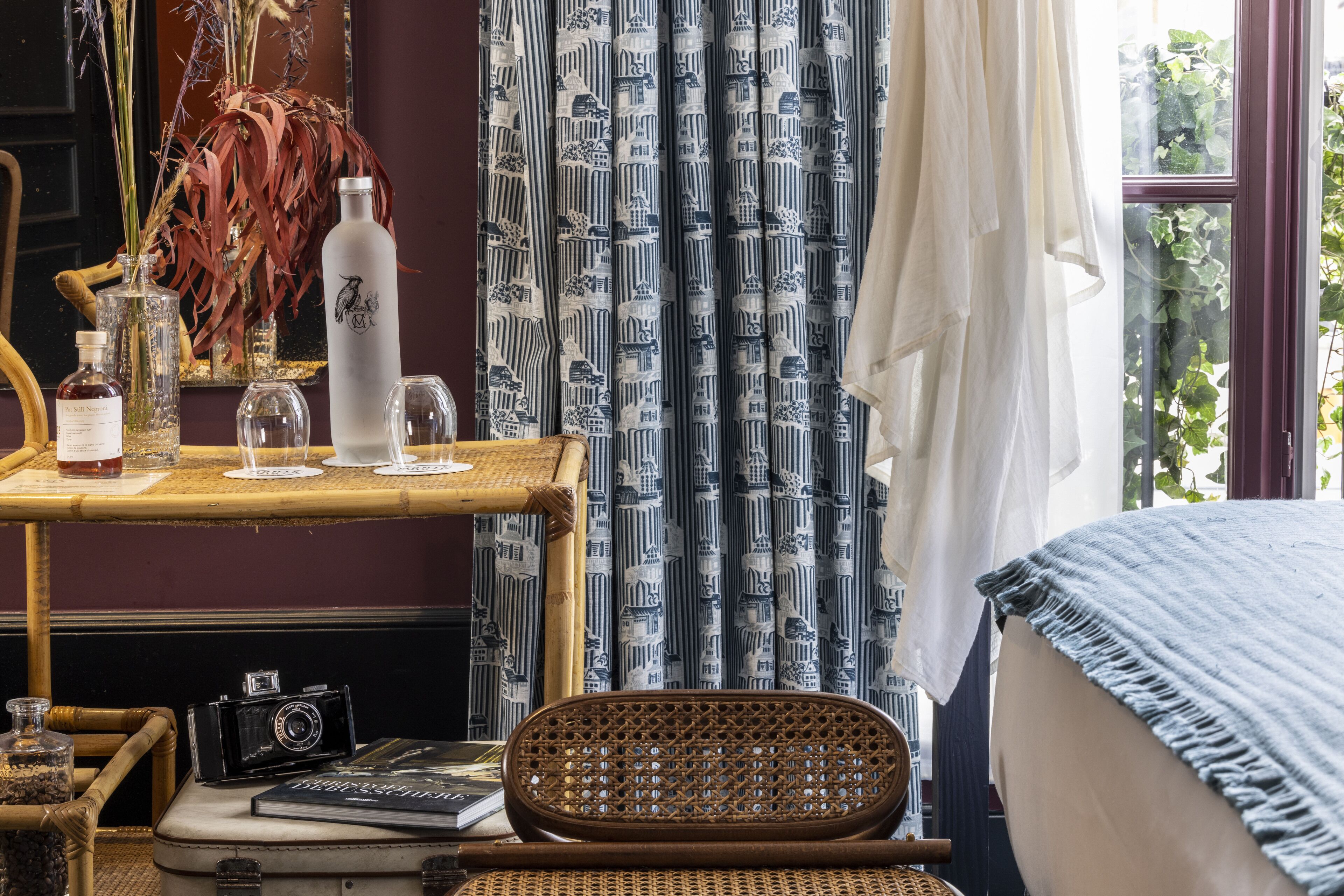 Una habitación con una cama, una silla de mimbre, una mesa con botellas, una cámara y cortinas estampadas.