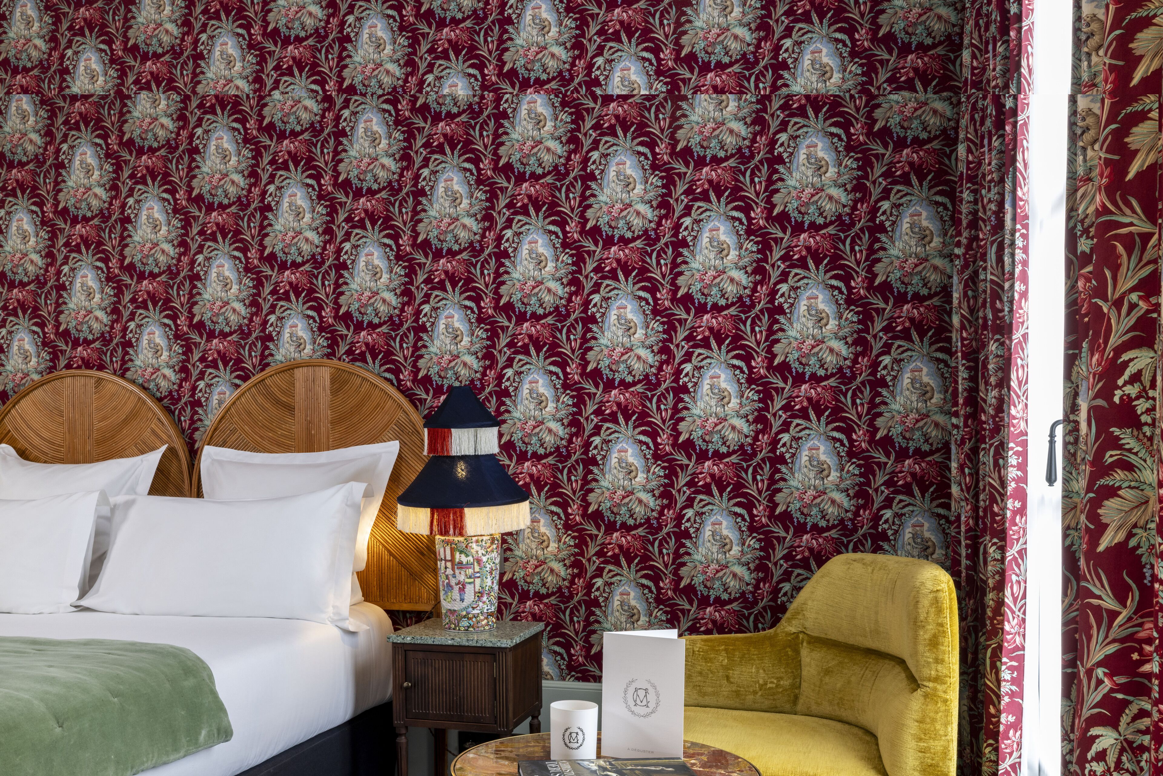 A hotel room with a large bed, a nightstand with a lamp, a chair, and a wall with a floral pattern.