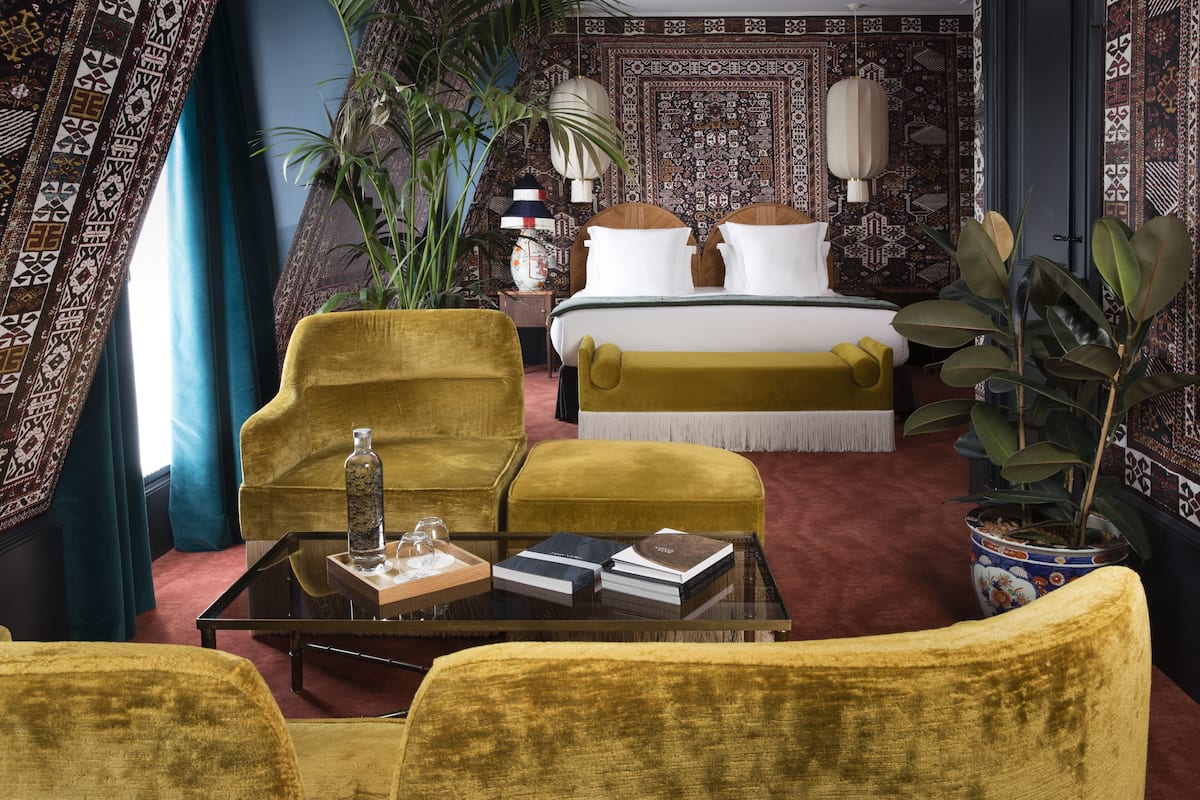 A room with a bed, two yellow armchairs, a glass coffee table with books and a bottle, and a large potted plant.