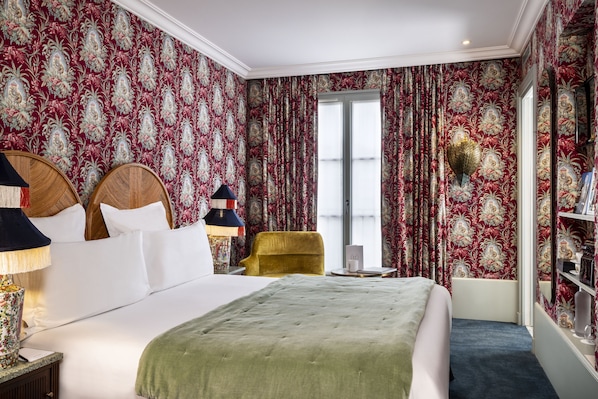 A hotel room with a large bed, a yellow armchair, a bookshelf, and floral wallpaper.