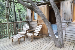 Eine Holzterrasse mit zwei Adirondack-Stühlen, eine Holzhütte mit einem Fenster und ein Baumstamm, der an der Hütte lehnt.