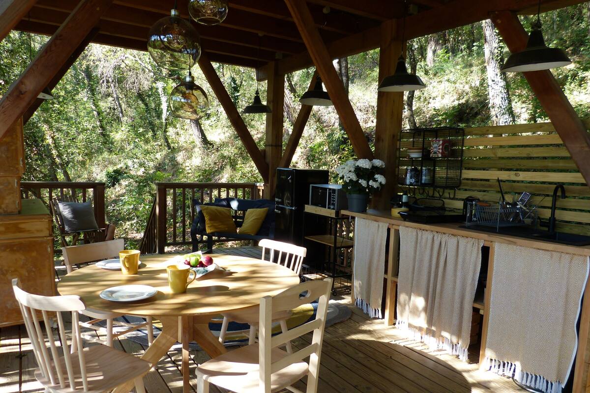 A wooden deck with a round table and chairs, a bar area with a sink, and a view of the surrounding forest.