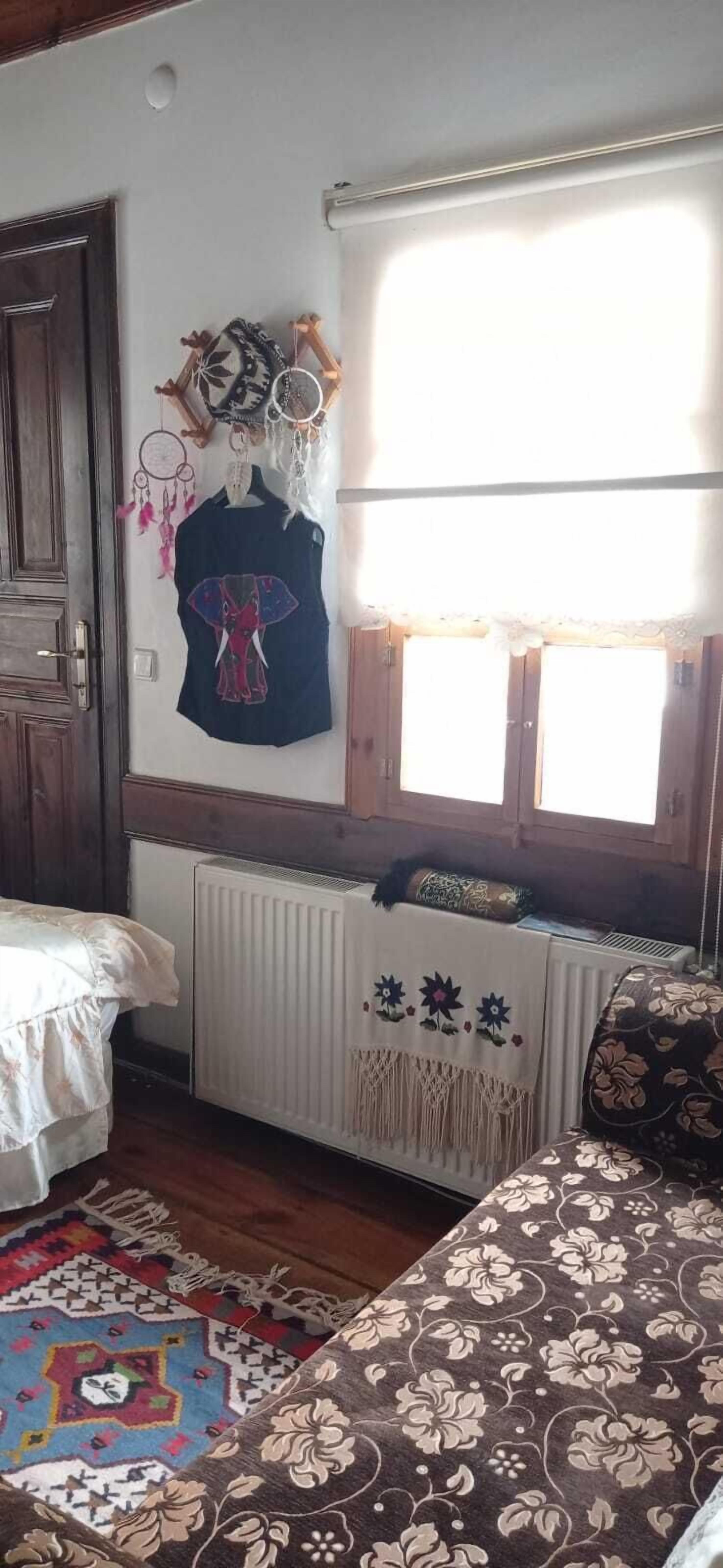 A room with a bed, a radiator, a window with curtains, a wall with dreamcatchers, and a patterned rug.