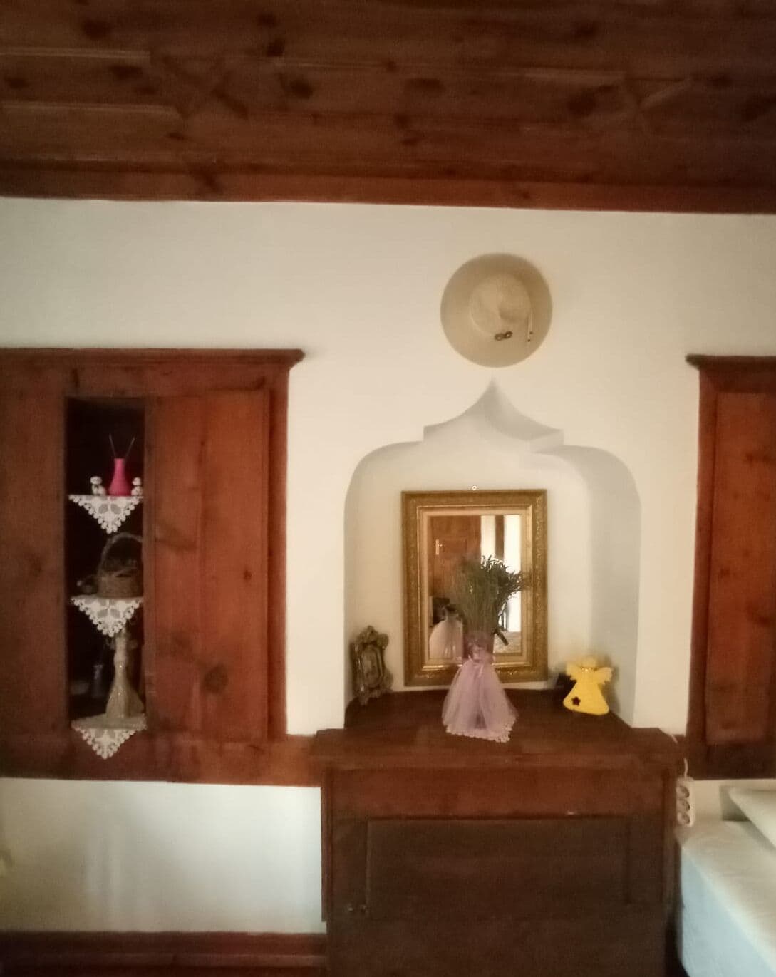Una habitación con techo de madera, un espejo con marco dorado, una mesita con un florero y una ventana con contraventanas de madera.