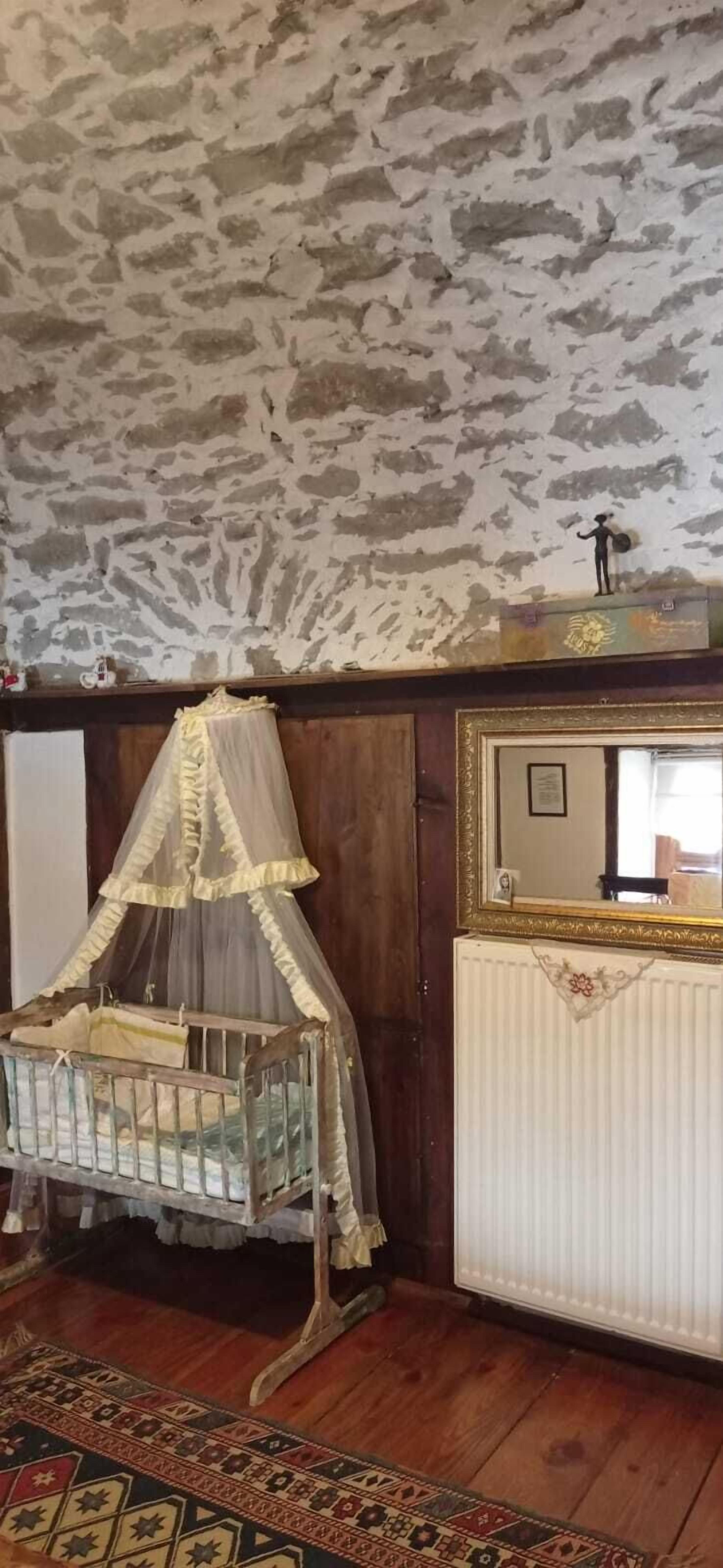 A room with a rustic stone ceiling, a wooden floor, a traditional crib with a canopy, a radiator, and a mirror with a decorative frame.
