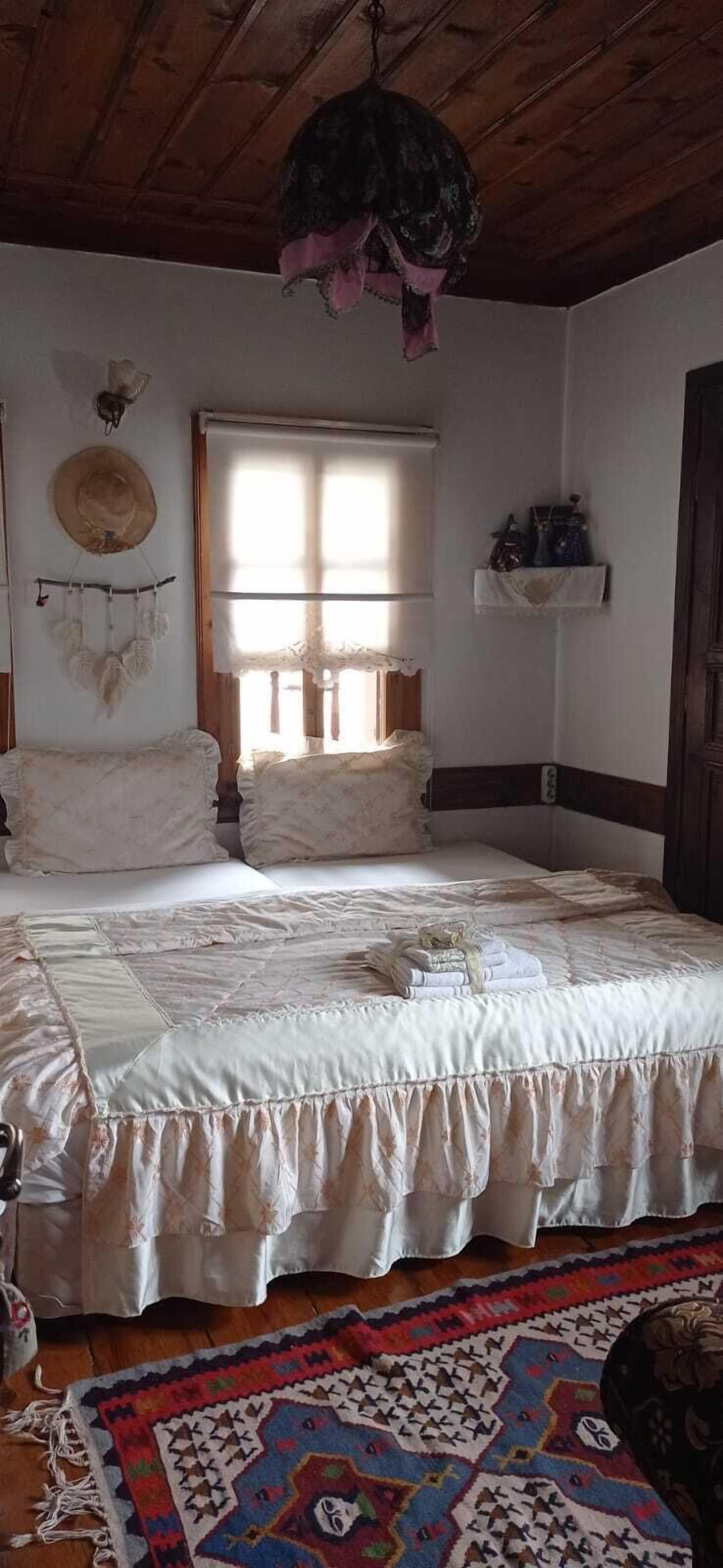 A bedroom with a bed, a hanging light fixture, a dreamcatcher, a window with blinds, and a colorful rug.