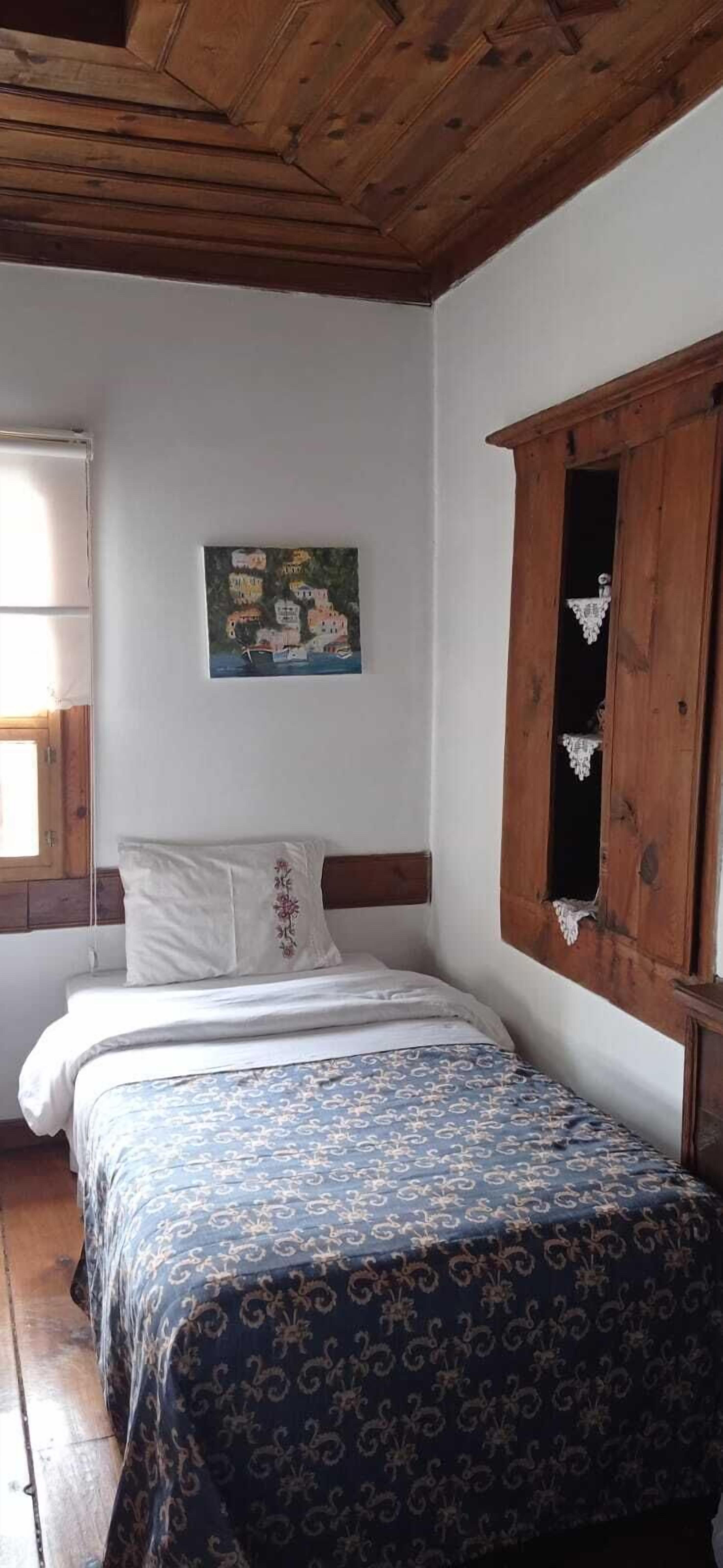 A bedroom with a wooden ceiling, a bed with patterned bedding, a white wall with a painting, and a wooden cabinet with a mirror.