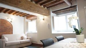 Une pièce avec des poutres apparentes, un mur en briques, un canapé blanc, une table à manger avec des chaises et une fenêtre ornée de fleurs blanches.