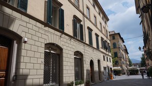 Un bâtiment historique doté d’arcades et de volets verts, comprenant un espace public avec une fontaine et des sièges en plein air.