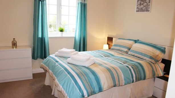 Una cama bien hecha con ropa de cama a rayas, una mesita de noche con lámpara, un aparador y una ventana con cortinas.