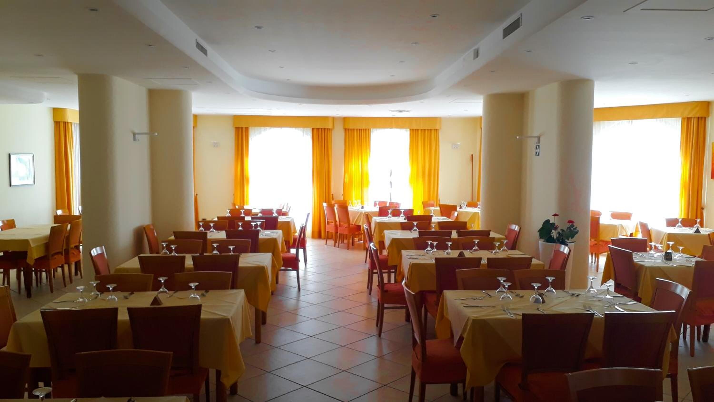 A large dining hall with multiple tables and chairs, arranged in rows, with yellow curtains and a tiled floor.