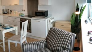 Een moderne keuken met witte kasten, een roestvrijstalen afzuigkap en een magnetron. Op de voorgrond staat een gestreepte fauteuil en een eettafel met stoelen.