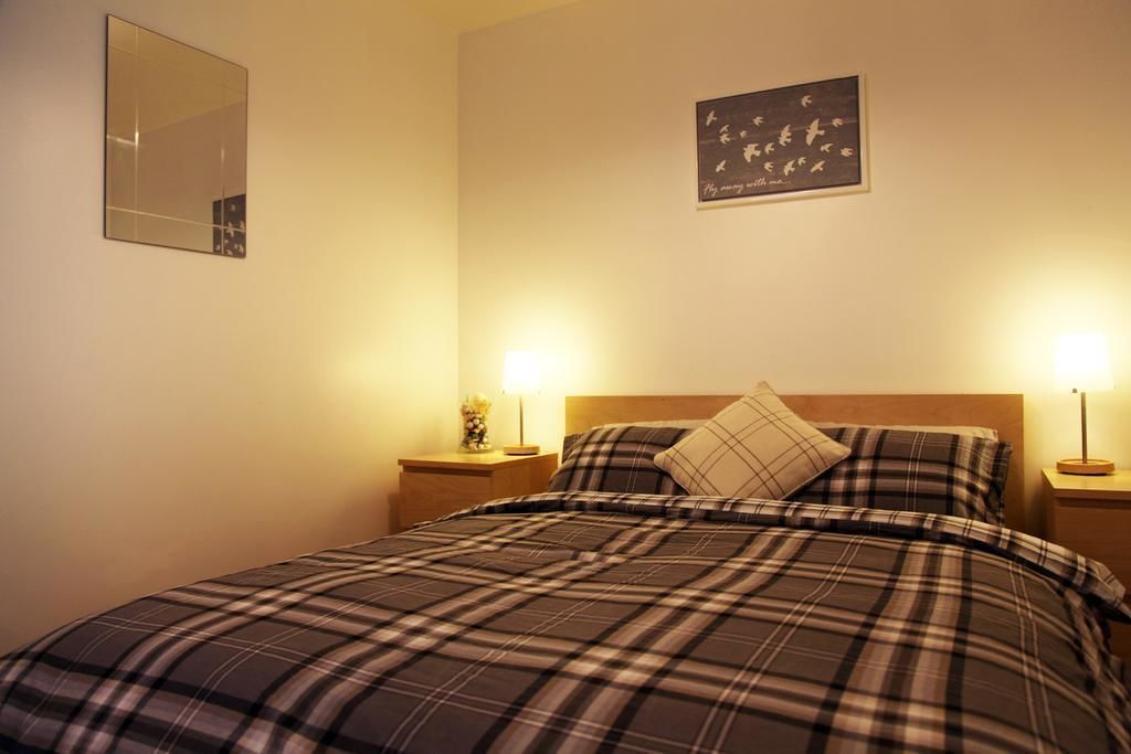 A neatly made bed with a plaid bedspread, two bedside lamps, a nightstand with a vase, and a framed picture on the wall.