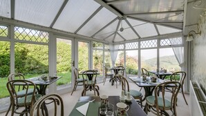 A conservatory with tables and chairs, large windows, and a view of greenery. - Cleeve Hill Hotel (Cheltenham)