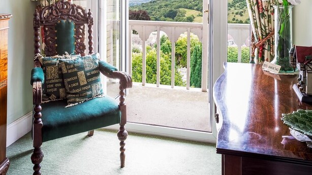 A room with a view of a green landscape, a wooden chair with a cushion, a table with a vase of flowers, and floral curtains. - Cleeve Hill Hotel (Cheltenham)