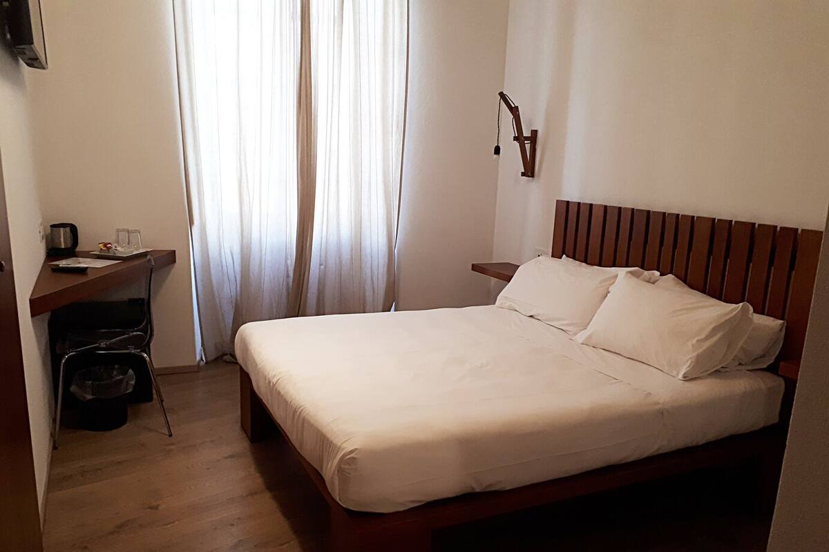 Habitación de hotel con cama de madera, un escritorio con silla, un cesto de basura pequeño y una lámpara fijada a la pared.