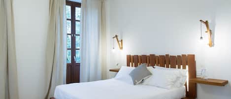 Un dormitorio con cama de madera, ropa de cama blanca, cabecera de madera, una mesita de noche y una ventana con cortinas.