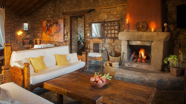 A rustic room with a stone fireplace, a white sofa with yellow cushions, a wooden table with a fruit bowl, and a stone wall with a painting. - Finca El Cabezo (Villamiel)