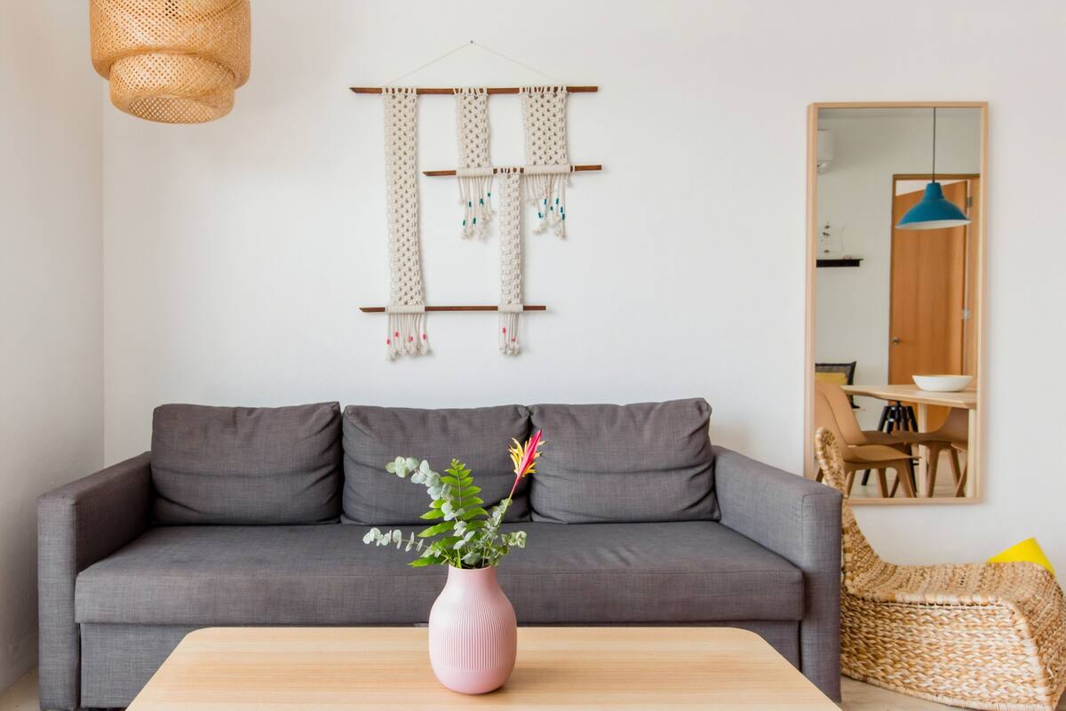 A modern living room with a grey sofa, a pink vase with flowers, a woven chair, and a macrame wall hanging.
