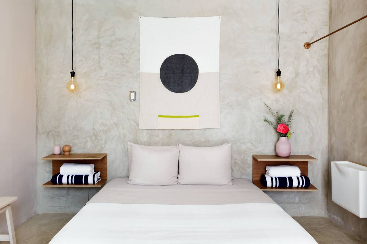 A modern bedroom with a large bed, two bedside tables with towels, a wall-mounted artwork, and two hanging light bulbs.