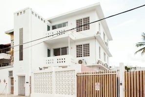 A white two-story building with a gated entrance, a balcony on the upper floor, and a visible security camera. - TROPICALIA by DW (San Juan)