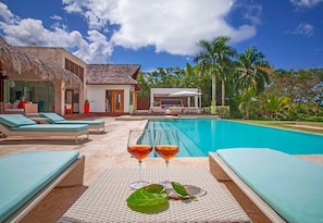 A poolside area with lounge chairs, a pool, and a building with a thatched roof. - Villa Perla by Unlimted Luxury Villas (La Romana)