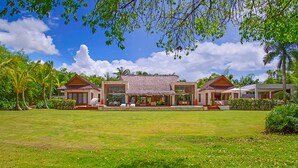 A large, two-story building with a thatched roof, surrounded by lush greenery and palm trees. - Villa Perla by Unlimted Luxury Villas (La Romana)