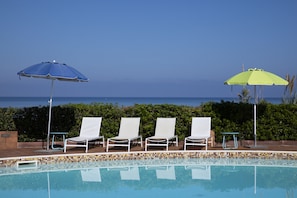 A poolside area with lounge chairs and umbrellas, a tiled pool, and a view of the ocean.