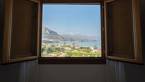 A window with wooden frames and white curtains, offering a view of a coastal town with mountains and a body of water. - Riva Sea Apartments (Castellammare del Golfo)
