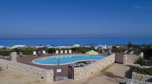 Una piscina con un muro de piedra y sillones de descanso, rodeada de sombrillas, con vista al océano.