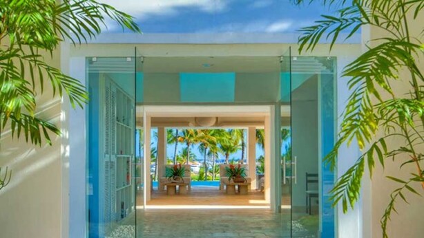 A modern hotel entrance with glass doors, a pathway leading to a seating area with a view of palm trees and a pool. - Casa Feliz by Unlimited Luxury Villas (La Romana)