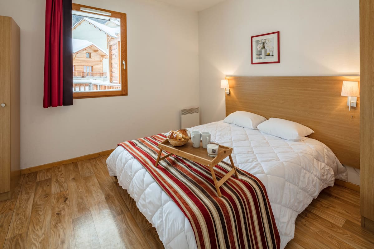 Ein Hotelzimmer mit Holzboden, einem Bett mit gestreifter Decke, einem Nachttisch mit Frühstücksangebot, einem Fenster mit rotem Vorhang und einem an der Wand hängenden Bild.