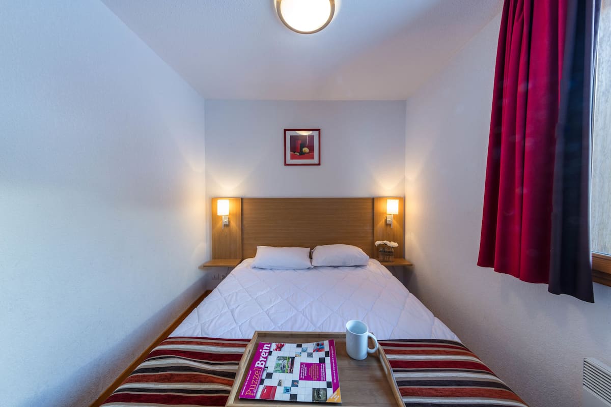 Ein Hotelzimmer mit Bett, einem Tablett mit einer Zeitschrift und einem Fenster mit roten Vorhängen.