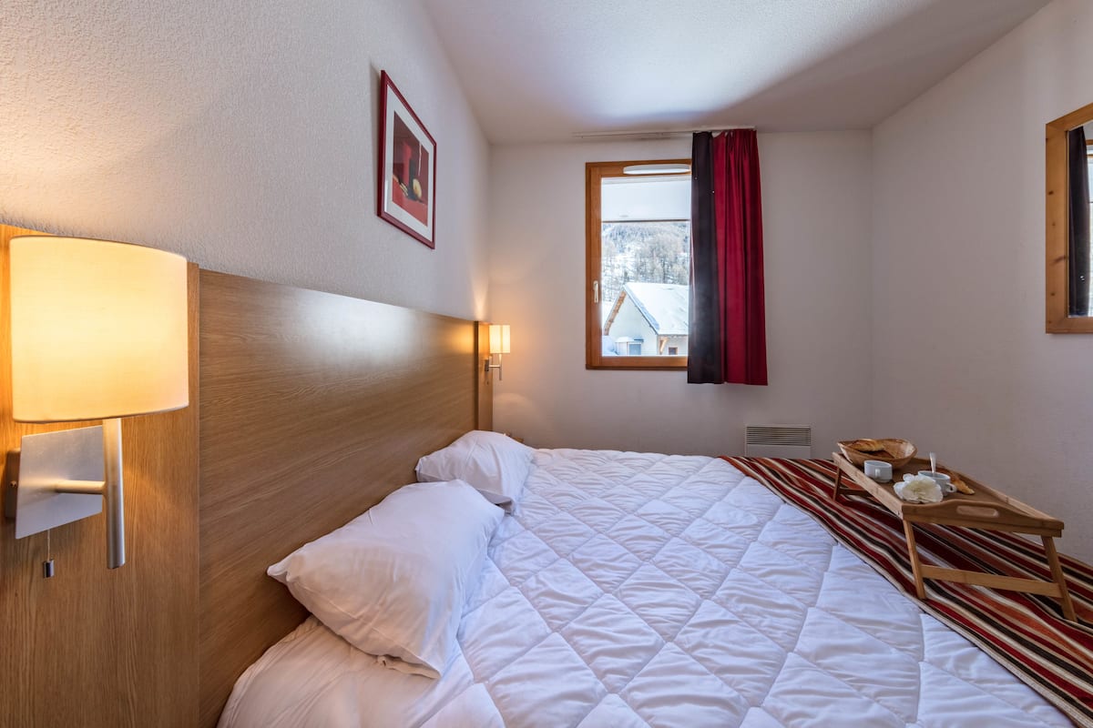 Ein ordentlich bezogenes Bett mit weißen Leinen, ein hölzernes Kopfteil, ein Nachttisch mit Lampe, ein gerahmtes Bild an der Wand und ein Fenster mit rotem Vorhang.