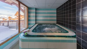 A hot tub with a view of snow-covered mountains, a tiled wall, and a window with a wooden balcony. - Noemys Les Balcons de Bois Méan (Les Orres)