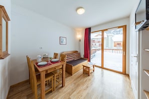 A room with a wooden dining table, chairs, a bench with cushions, a small side table, and a sliding glass door leading to a balcony. - Noemys Les Balcons de Bois Méan (Les Orres)